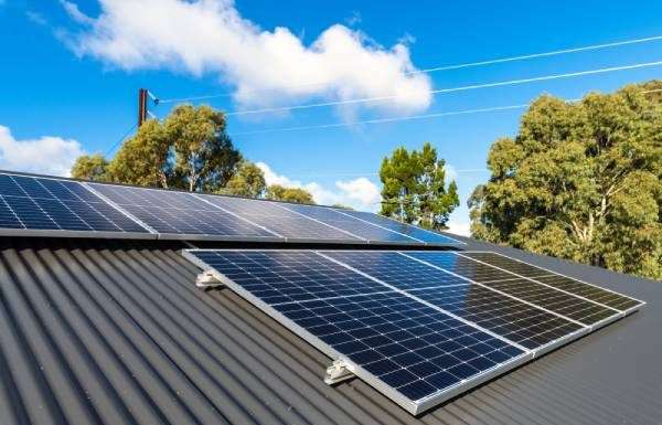 A metal roof with rows of solar panels on a partly-cloudy day. Trees and telephone wires are seen in the background.