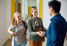 A teenage girl wearing a backpack smiles as her mother shakes hands with her teacher in the hallway of the school.