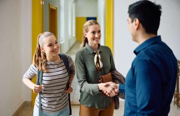 A teenage girl wearing a backpack smiles as her mother shakes hands with her teacher in the hallway of the school.