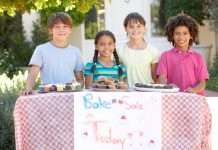 A group of children standing outside behind a bake sale table and smiling. There are muffins and cupcakes on the table.