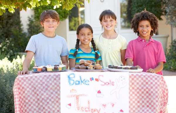 A group of children standing outside behind a bake sale table and smiling. There are muffins and cupcakes on the table.