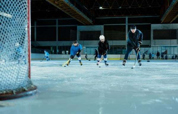 A hockey practice on an indoor rink. Players practice in the background while two young players skate toward the goal next to their coach.