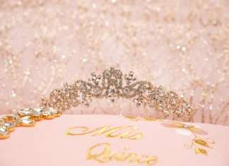 A sparkly quinceañera tiara rests on a pink pillow that has ‘Mis Quince’ embroidered on it in gold thread.