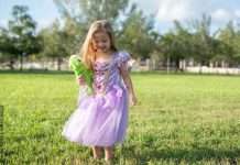 A little girl wearing a purple princess gown. She is standing in a field holding a stuffed animal of a chameleon.