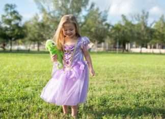 A little girl wearing a purple princess gown. She is standing in a field holding a stuffed animal of a chameleon.