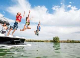 Two boys and a girl wearing life jackets and jumping off the back of a boat into a lake. Their mother is watching from the boat.