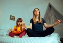 A young mother unwinds by practicing her daily yoga meditations with her daughter on the daughter's bed.