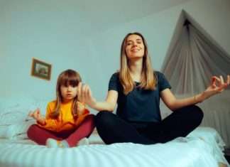 A young mother unwinds by practicing her daily yoga meditations with her daughter on the daughter's bed.