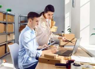 Two people working at a desk with a laptop on it. Several cardboard boxes neatly organized on shelves fill the background.