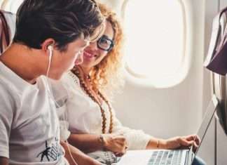 A woman sits by the window of an airplane with a laptop and looks over to a teen boy with headphones.