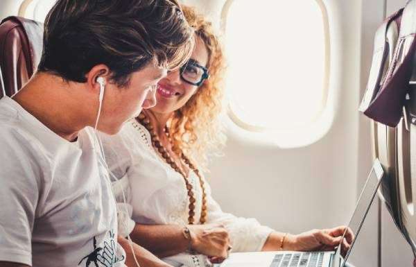 A woman sits by the window of an airplane with a laptop and looks over to a teen boy with headphones.