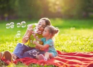 A young woman in a neon green shirt sitting on a red blanket with a little girl in polka dots, blowing bubbles together.