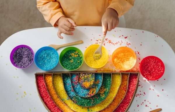 A child plays with red, orange, yellow, green, blue, and purple rice and places it in a rainbow-shaped container.