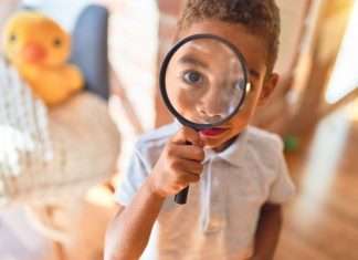 A child holds up and looks through a magnifying glass, which enlarges his eye. A duck toy sits behind him.