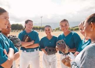 A female softball coach talks to her players in uniform in a circle. The players are holding their gloves and smiling.