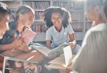 A group of kids sit on the floor in a circle, each holding a different book and smiling while facing one another.