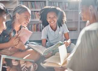 A group of kids sit on the floor in a circle, each holding a different book and smiling while facing one another.