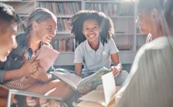 A group of kids sit on the floor in a circle, each holding a different book and smiling while facing one another.