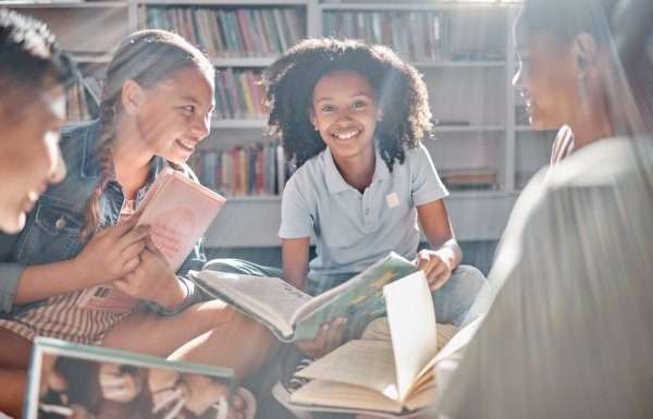A group of kids sit on the floor in a circle, each holding a different book and smiling while facing one another.