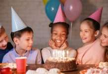 Small children in colorful, polka-dot party hats gathered around a birthday cake with multiple lit candles.