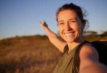 A young, smiling woman with a backpack taking a selfie outdoors and gesturing toward a grassy field.