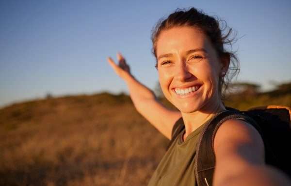 A young, smiling woman with a backpack taking a selfie outdoors and gesturing toward a grassy field.