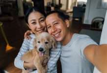A smiling man and woman holding a happy dog in a warmly lit room with wooden floors and cozy furniture.