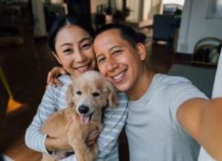 A smiling man and woman holding a happy dog in a warmly lit room with wooden floors and cozy furniture.
