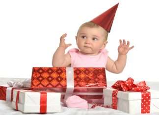 A baby girl wearing a pink set has a red hat on and surrounded by multiple red and white gift boxes.