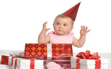 A baby girl wearing a pink set has a red hat on and surrounded by multiple red and white gift boxes.