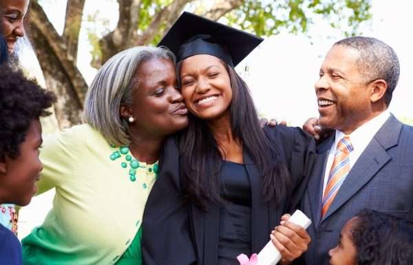 5 Surprises You Can Give Your Kids at Their Graduation A young female in a black graduation gown and hat is being embraced by two parents in formal attire. Two children look on.