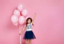 A happy and fashionable young girl is posing with pastel pink birthday balloons on a pink background.