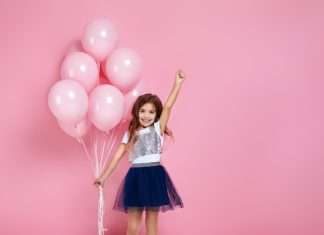 A happy and fashionable young girl is posing with pastel pink birthday balloons on a pink background.