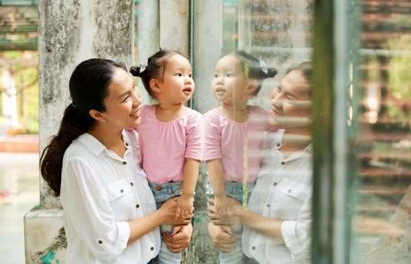A mother holds up her small child so that she can look through a glass wall at a zoo. Both are smiling.