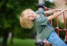 A smiling child, dressed in a green t-shirt and jeans, playfully hanging off of a rope section on a playground.
