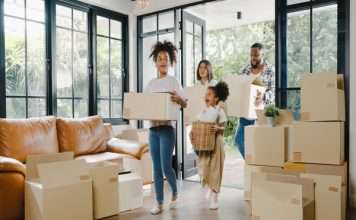 A family of two adults and two children is walking into a home with large windows and stacks of moving boxes.
