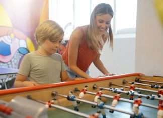 A woman and a young boy smile while playing foosball together at a table in a bright indoor recreation area.